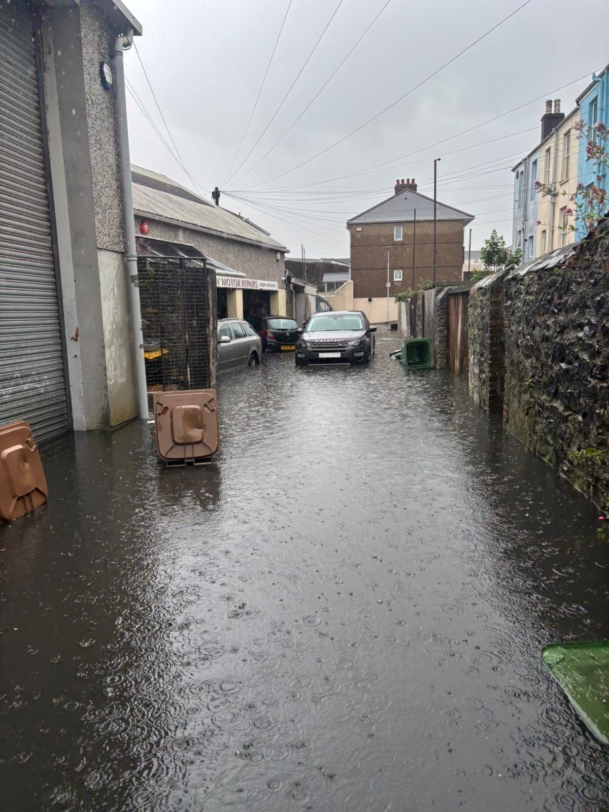 An urban lane is flooded with water, there are cars in the floodwater and back garden gates open onto the laneway.  To the left of the picture is a garage, with signage above the large open doors.  