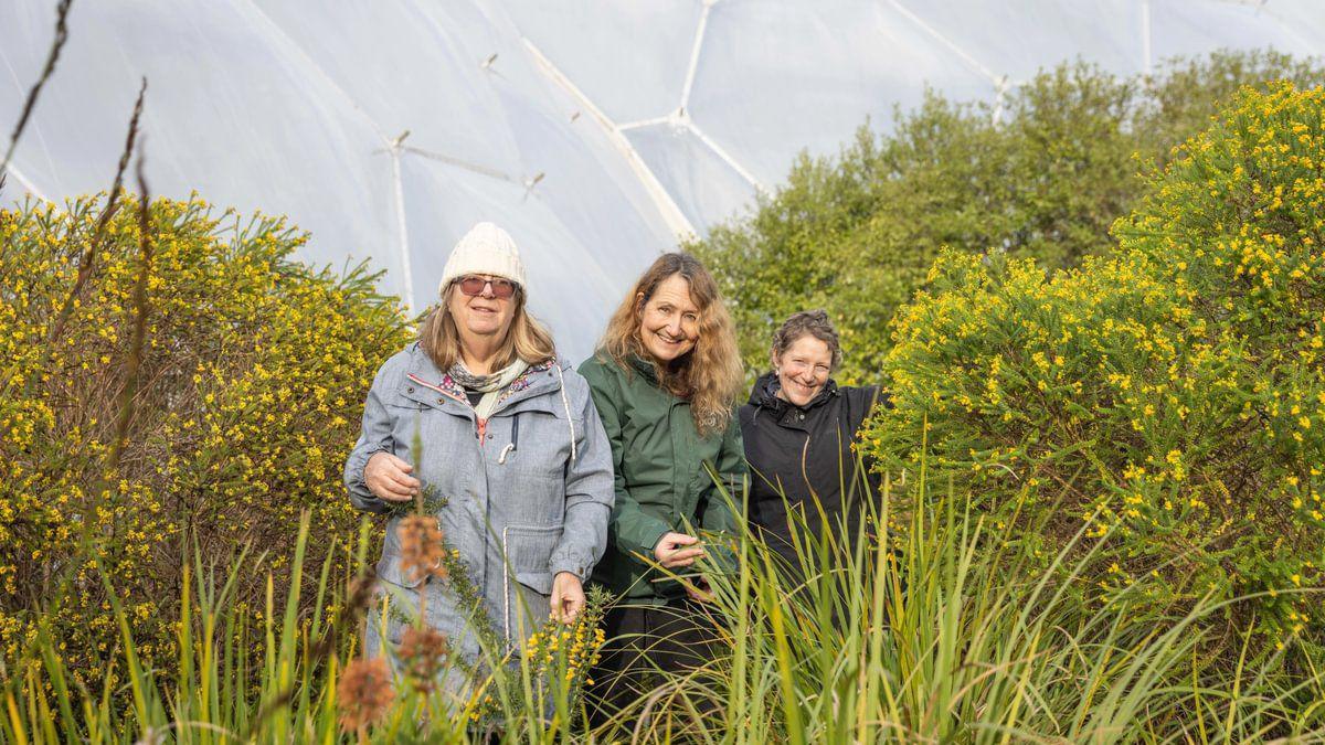 Three women standing in a line next to greenery. Behind them is the Eden Project's while biome. 