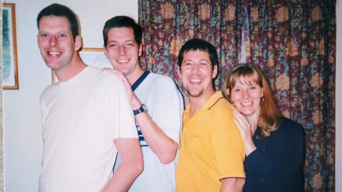Tim, Andy, Richard and Sue stand in height order sometime in the 1990s. Tim and Andy are both wearing white shirts while Richard is in a bright yellow shirt and wears glasses. Sue has shoulder length blonde hair and a dark blue top. They are all smiling widely and there is a red and yellow floral patterned curtain in the background.