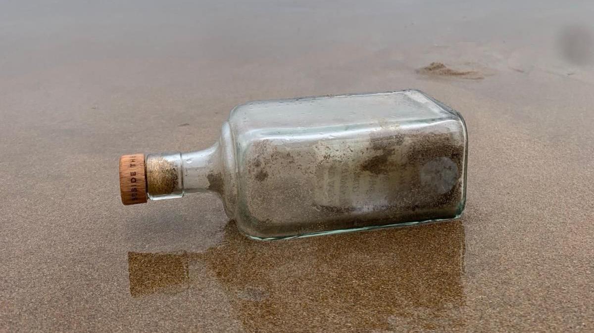 A bottle with a cork. There are ashes inside the bottle and a note. It is lying on its side in the sand. 