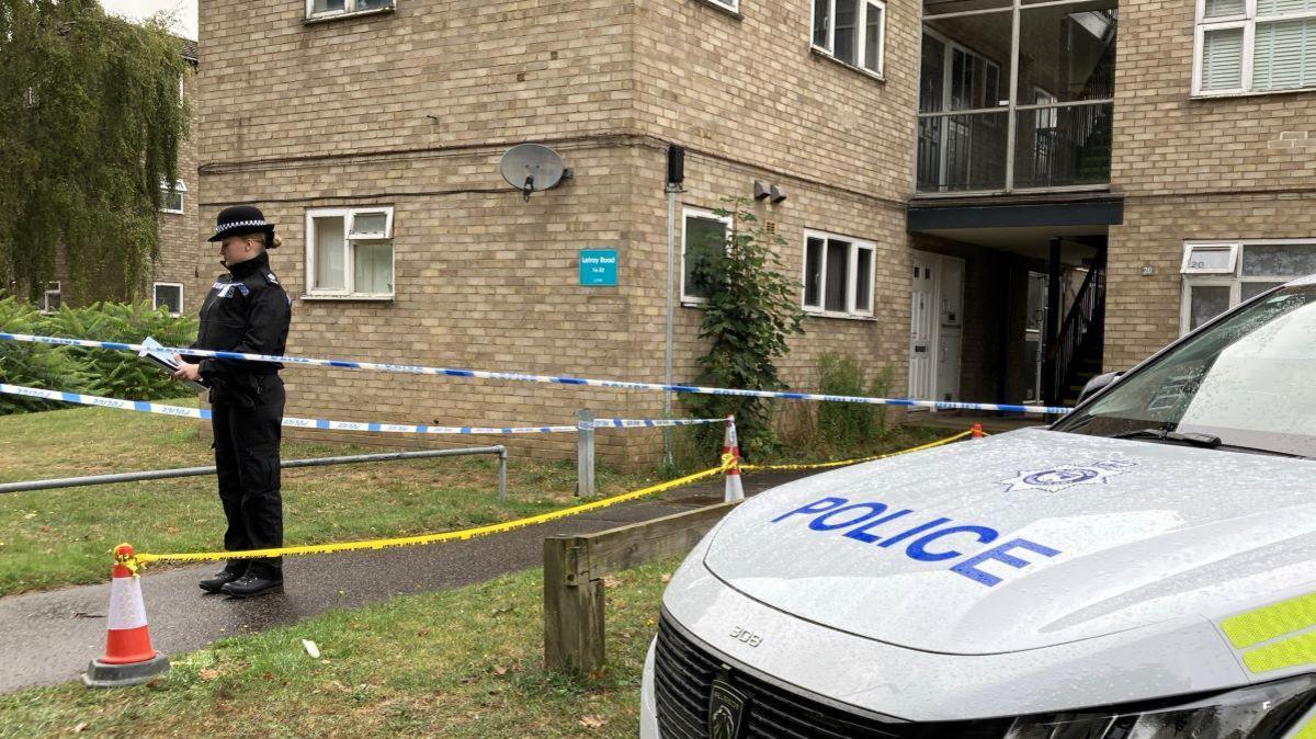 A police officer stood inside a police cordon outside a block of flats, with the bonnet of a police car in the foreground