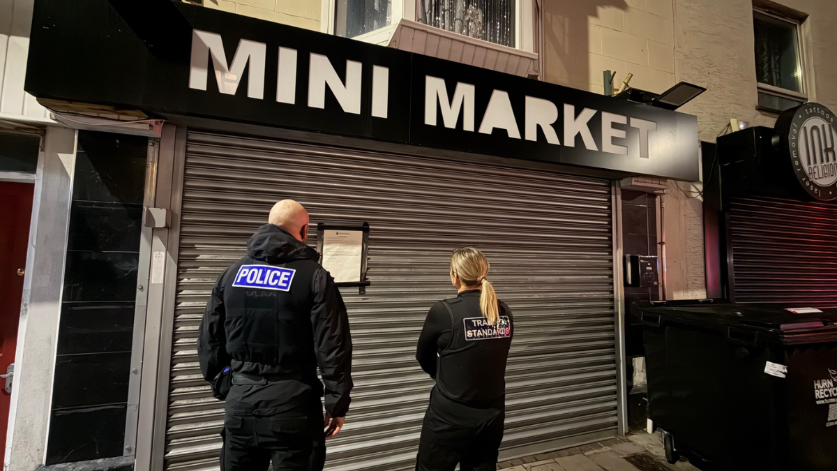 A police officer and trading standards officer standing outside a shop with a grey shutter. There is a black sign with the words 'Mini Market' in white. The shutter is down and there is a sign on the left. 
