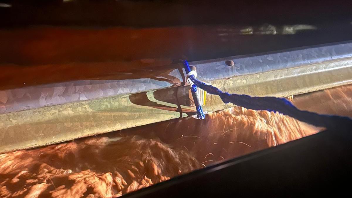 A view from a car window, showing a rope attached to a metal barrier while brown water flows beneath, just under the height of the barrier.