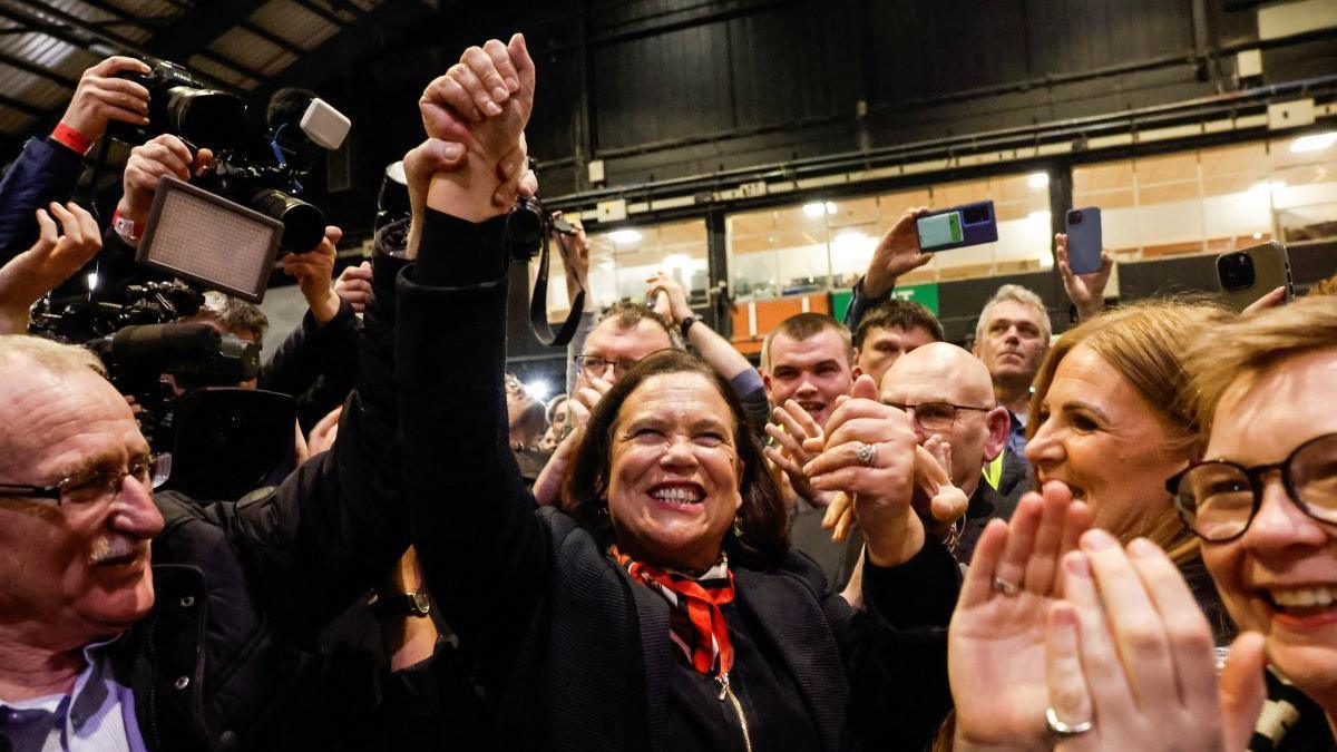 Mary Lou McDonald holding her hand up in the air as she celebrates. She has shoulder length brown hair and is wearing a black blazer with an orange and black scarf.