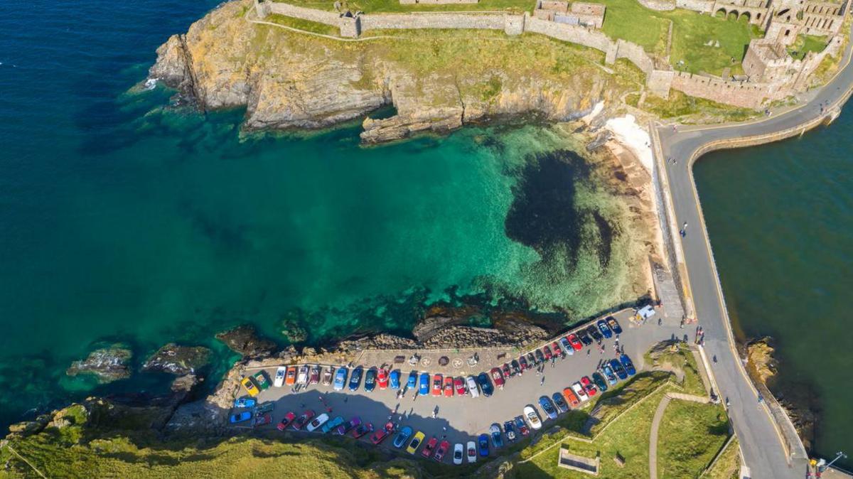 An aerial view of the car park at Fenella Beach in Peel with dozens of colourful cars lined up near to the sea and Peel Castle.