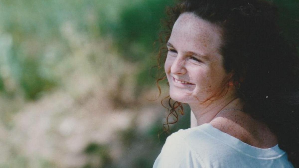 A smiling image of Sammy Young pictured when she was in her 30s. She has long brown curly hair, freckles on her forehead and is looking over her shoulder, smiling. She is wearing a white top with a low back. The background is out of focus.