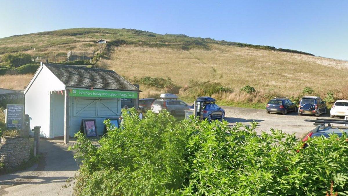 Google image of National Trust's Baggy Point car park. A small hut is at the entrance of the car park. A sign states 'join here today and support Baggy Point'. A number of cars are parked in the car park which is surrounded by fields. 