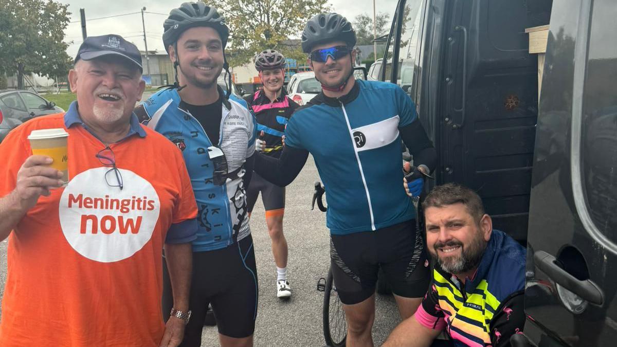 Four cyclists and one man pictured in a bright orange 'Meningitis Now' t-shirt smile while standing next to a van parked in a car park.