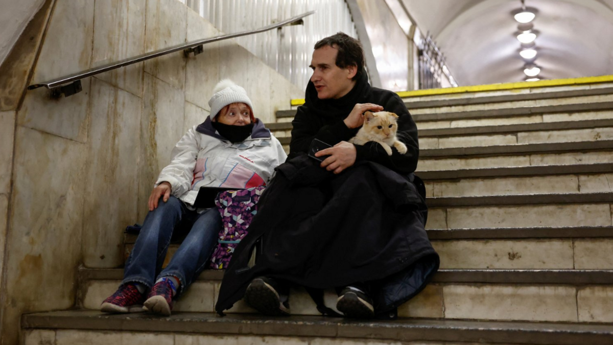 People take shelter inside a metro station during a Russian military attack. One man can be seen holding a cat while stroking its head while sitting next to a woman on the steps of a Metro station