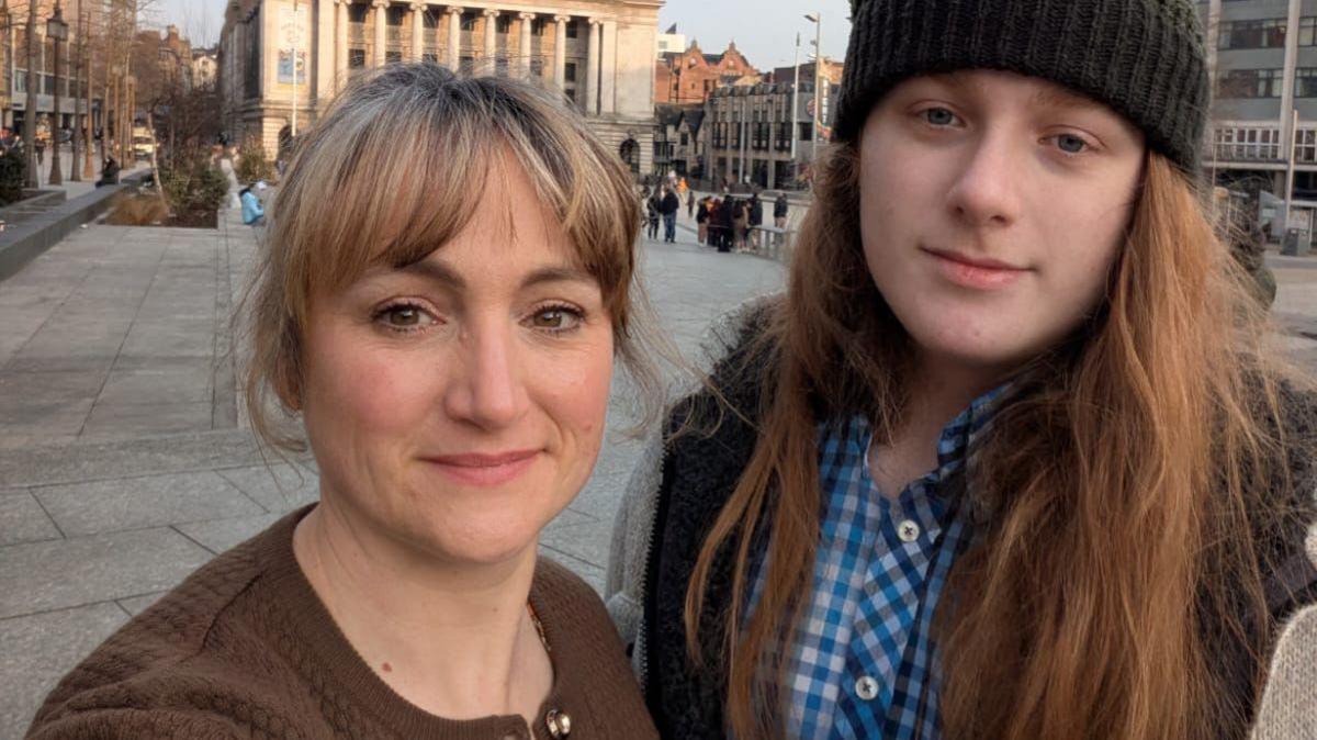 Woman with blonde hair and brown cardigan with teenage son in black beanie hat and long red hair, infront of grey town hall building in Nottingham