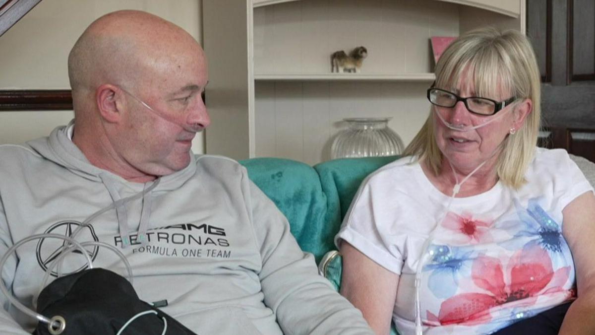 Mark Waud (left) and his sister Joanne Lovejoy-Waud are both sat next to each other on a sofa in the living room. The siblings both have transparent oxygen tubes attached to their face and feeding into their nostrils.