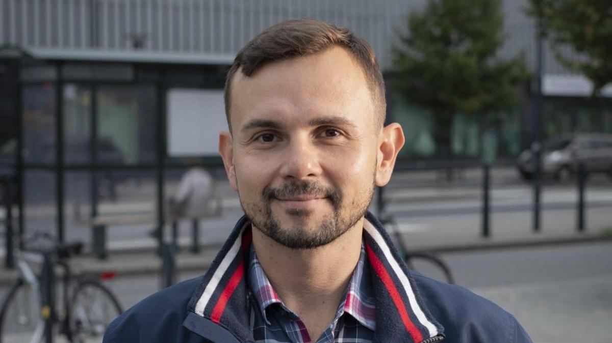 Dalibor Sentic is photographed stood outside on a street. A bike, bus stop and trees can be seen behind him. He has brown eyes, short brown hair, and a beard. He is wearing a plaid grey, red and white shirt and navy-blue jacket, which has a white and red stripe on the collar.