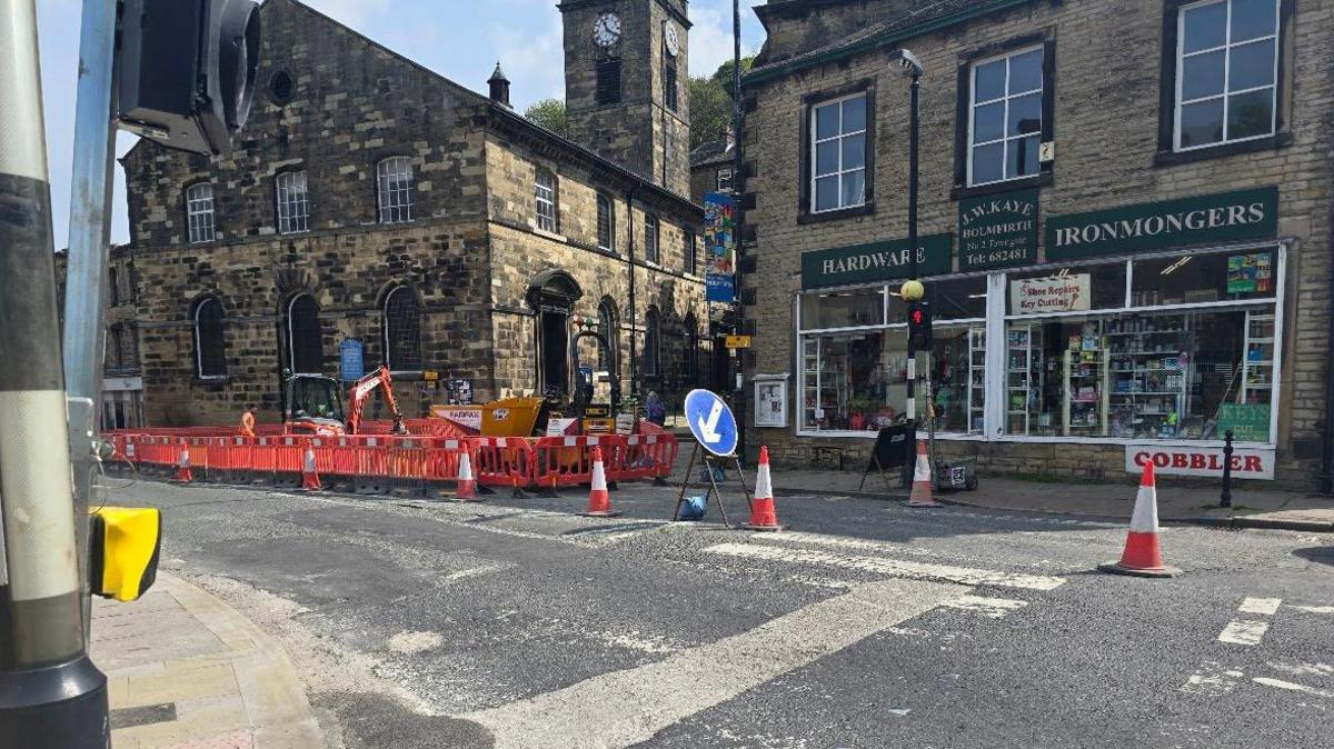 A road in Holmfirth with road words, traffic cones and a mini digger, in front of a row of shops.