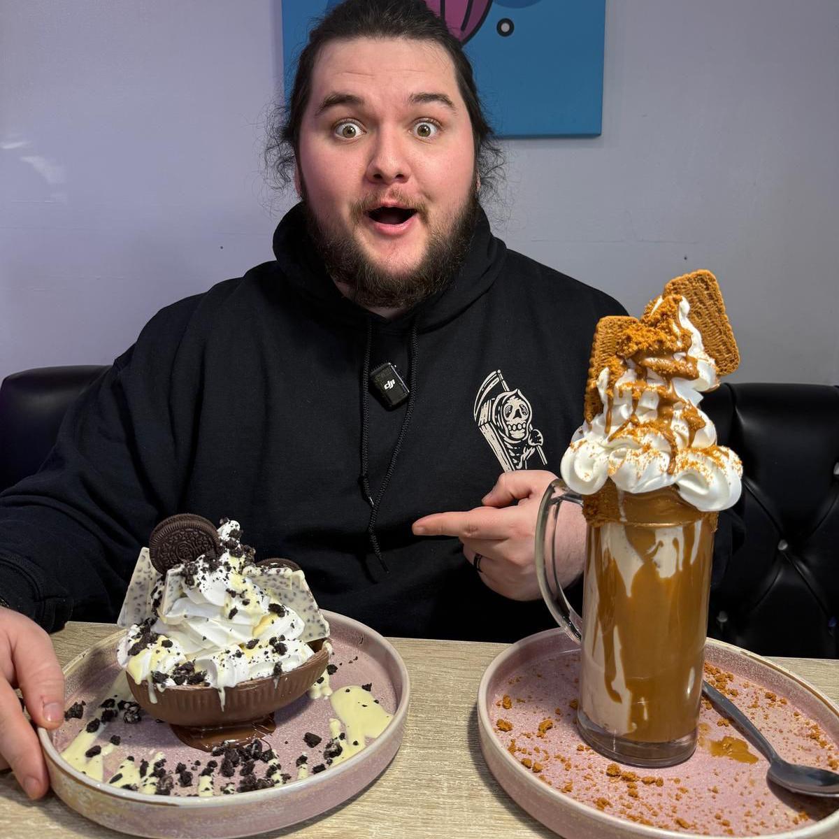 A man with a beard, dark hair and black hoodie, smiles as he points at one of two large desserts in front of him.