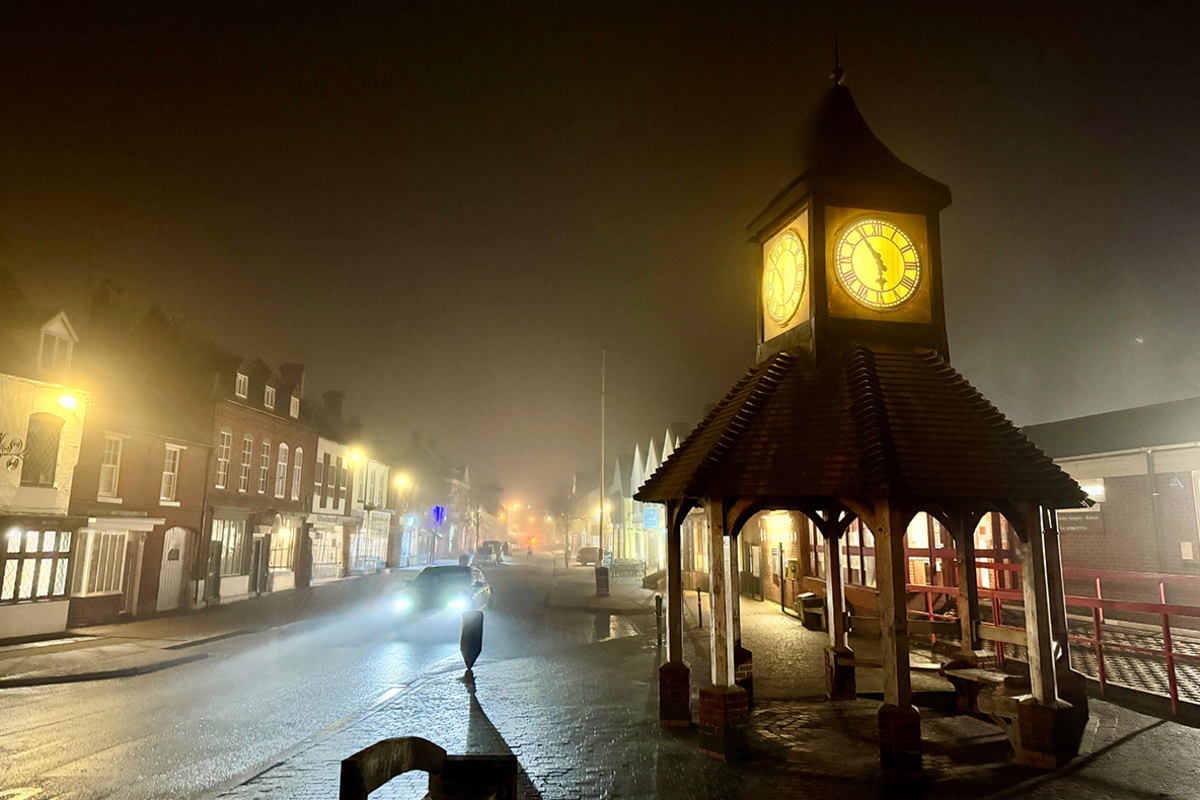 A covered bandstand with a illuminated clock as part of the structure stands in the foreground at the edge of a road junction. The sky is dark and lamp posts are lit up along the road as a car drives towards us with headlights on and buildings can be seen across the street.