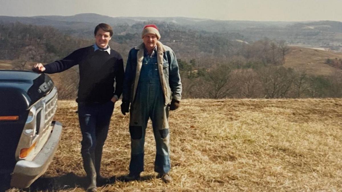 Tim Lobb seen as a younger man with his father Marshall Donohew. Mr Lobb is wearing boosts, jeans and a collared shirt. Mr Donohew is wearing denim overalls and a red beanie. Mr Lobb has his hand resting on an American truck. Behind them is open landscape.