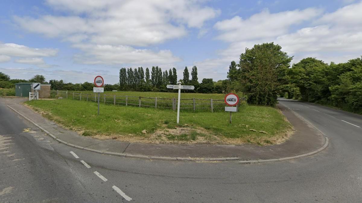 A view of a green field next to a road with trees. The sky is blue with white clouds.