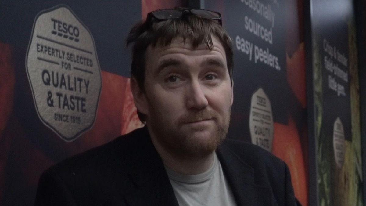 A man in a grey shirt and a black blazer. He has brown hair and a short, brown beard.
