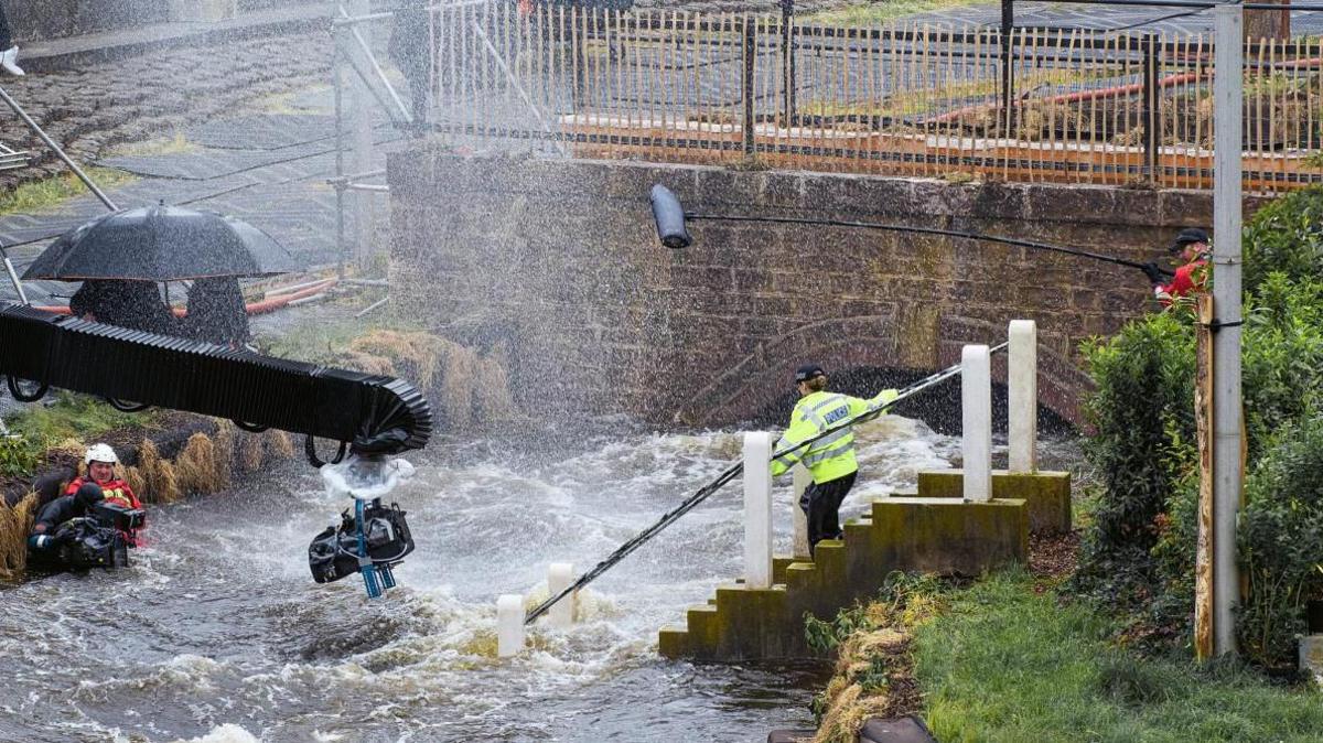 Filming equipment is rigged around a scene depicting high reiver flow under a bridge. An actor portraying a police officer can be seen running into the water