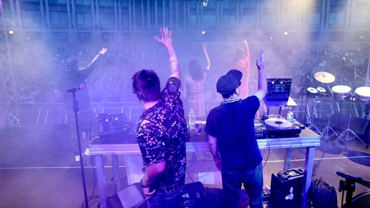 Two male DJs standing on stage at Bristol Harbour Festival with their arms in the air, hyping up the crowd below. The amphitheatre is full of a large crowd of people. The sky is dark and the image has an overall blue-purple hue from the stage lights.