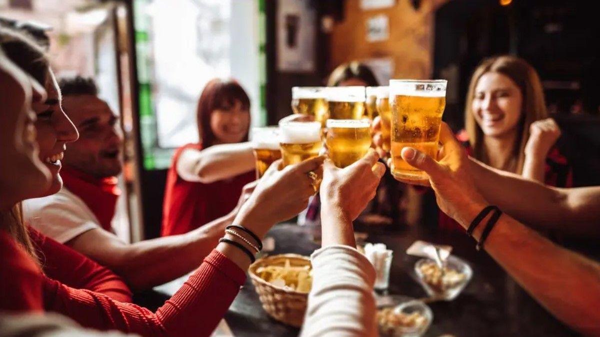 Eight people near a table are holding their beer glasses together in this generic image. Four of them are smiling and some faces are not visible.