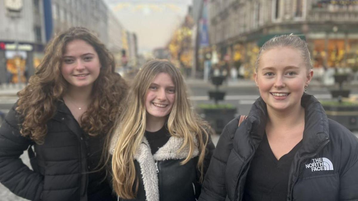 Three women aged 18, one with long curly brown hair and black jacket, another with blonde hair and black jacket and another with blonde hair wearing black jacket with black top. They are in Belfast city centre.