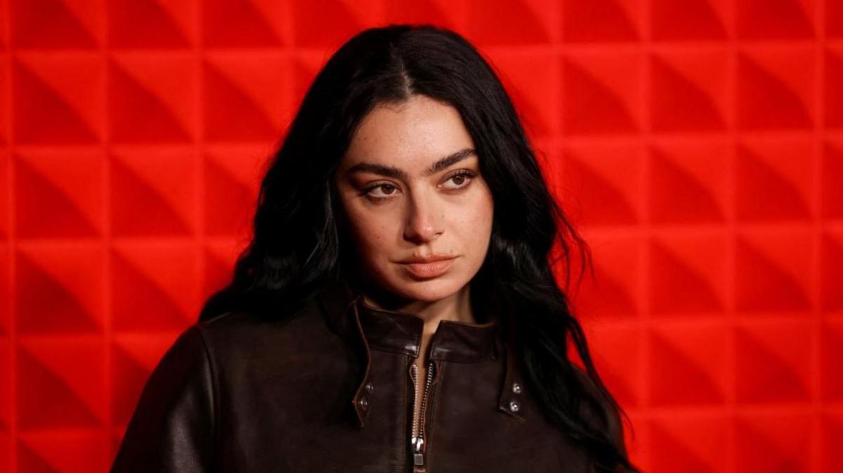 Charli XCX attends an event in London. She is wearing a brown leather jacket and is seen posing in front of a red wall. 