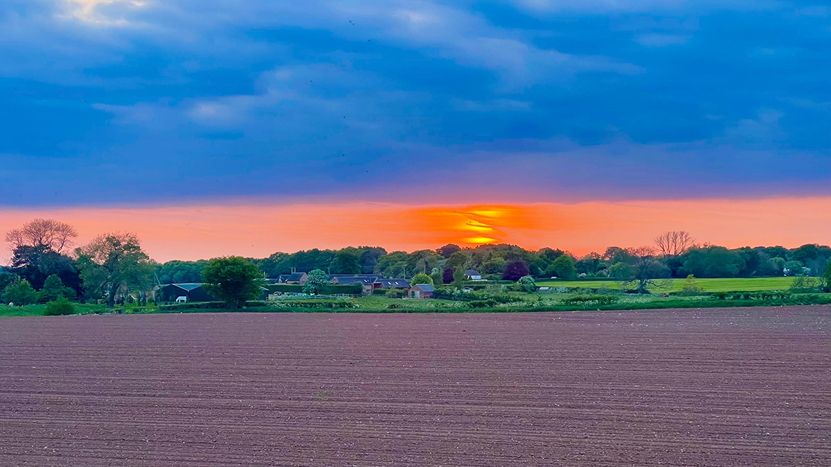 A ploughed field in the foreground with greenery beyond and a band of orange sky at  sunset or sunrise with darker clouds above