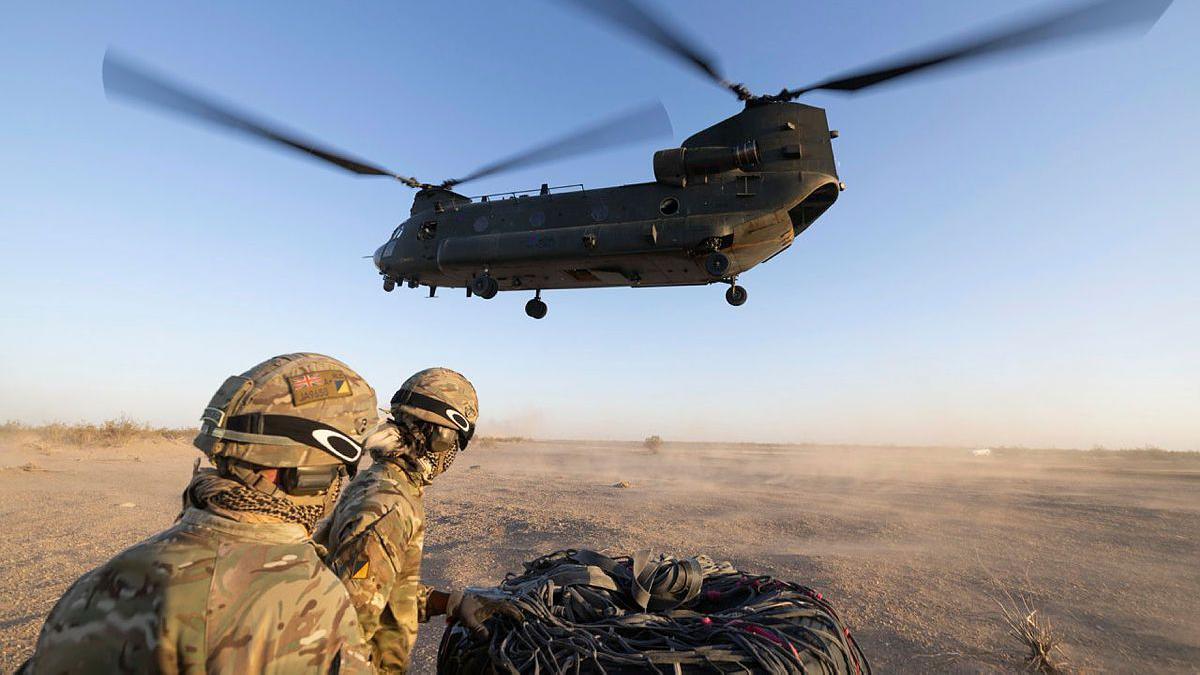 Two joint helicopter support squadron soldiers look away from camera while wearing camo gear and helmets, with a CH-47 Chinook mid air above them in a desert type environment. Image taken near El Centro USA in October.