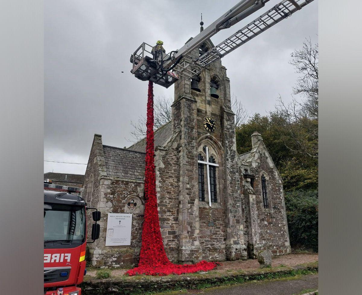 A firefighter in a cherry picker is seen draping the red poppy display on to a stone church building. Part of a fire engine is to the left of the image.