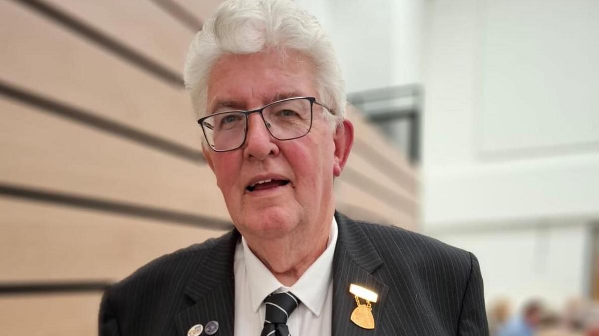 A man with glasses, in a black pinstriped shirt, white shirt and black tie looks at the camera. He is standing in a hall.