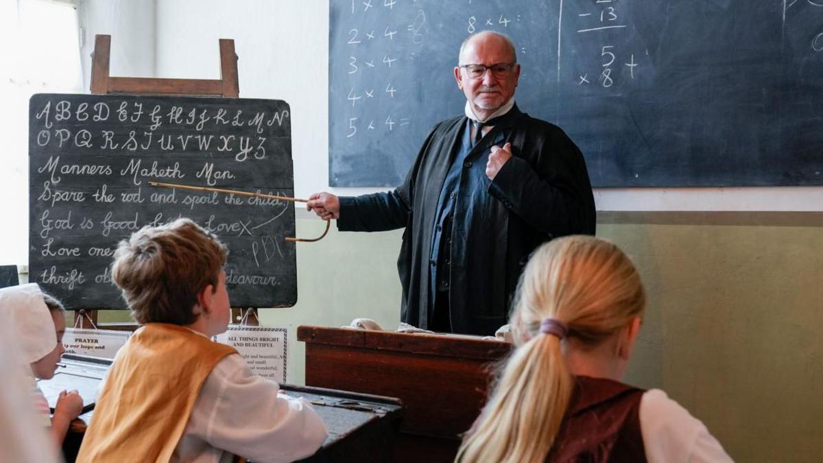 A man dressed in teachers robes points at blackboard with a cane.
The blackboard has old fashioned style writing in chalk on it.
Two children sit at wooden desks looking at him.