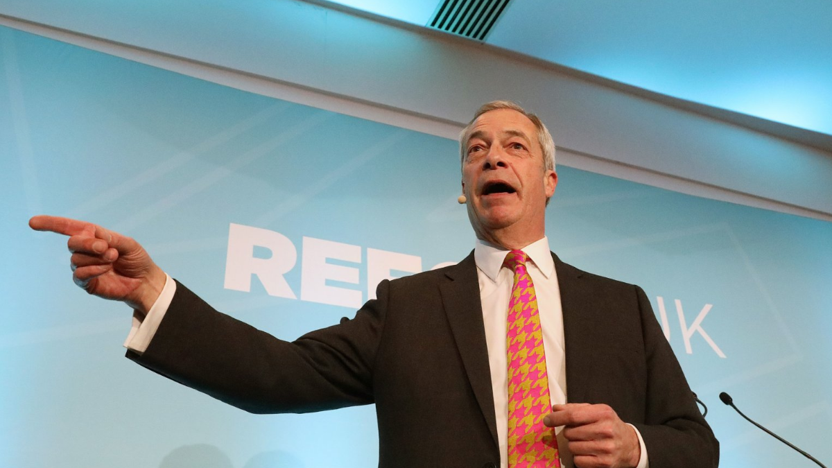 Nigel Farage, wearing a black suit with a white shirt and a fuschia pink and gold houndstooth tie, points during a speech.