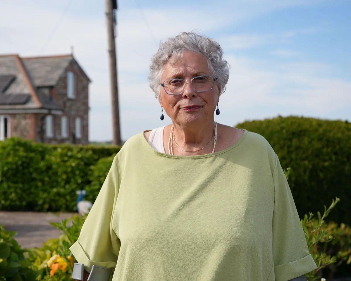 carole wyatt, a woman with short grey hair, wire frame glasses and a pale green tshirt, stands in front of a hedge and a house