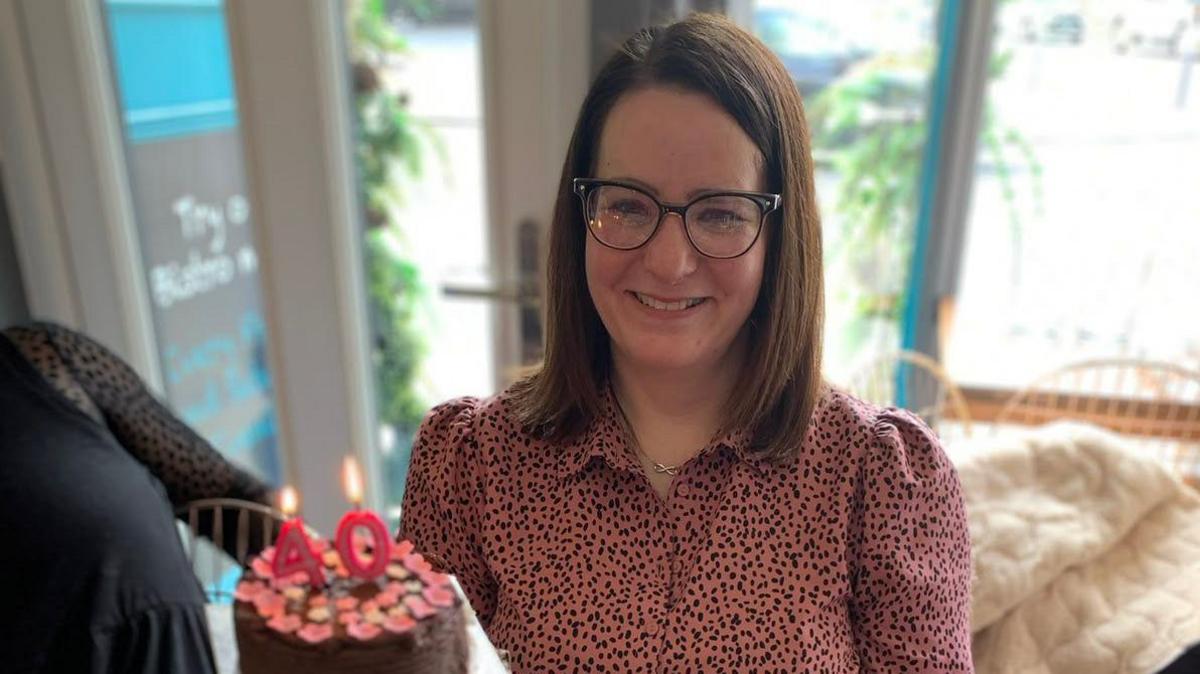 Anona Gauld is wearing a pink and black spotty shirt and black glasses. She is smiling and there is a 40th birthday cake in front of her. 