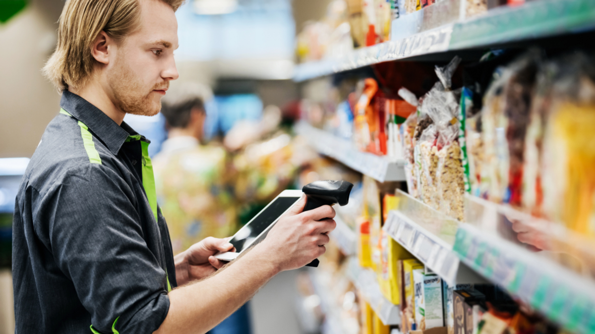 Young male supermarket worker, dressed in a dark shirt with green trimming, scans food on a shelf 