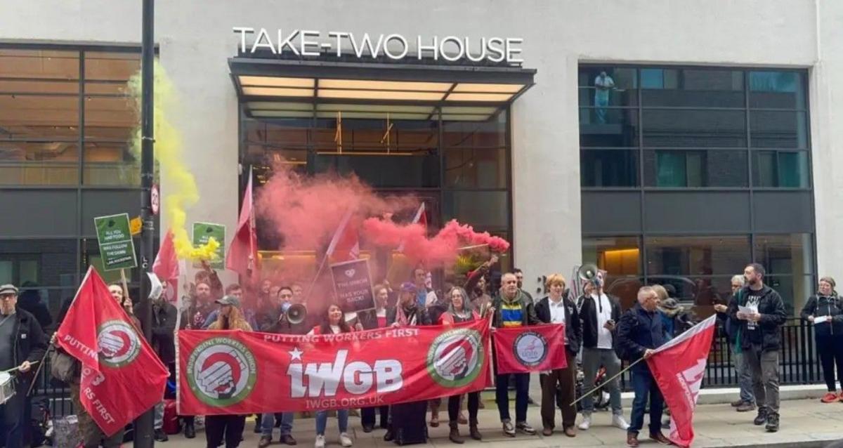 A crowd of people outside an office holding red and white banners which say IWGB and using red flares.
