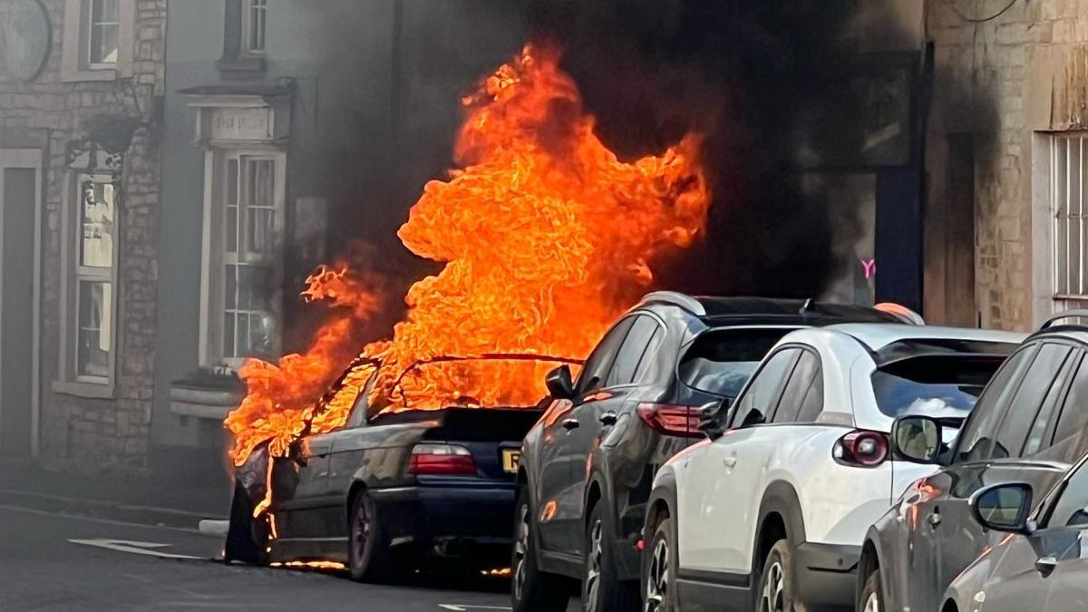 A black car on fire in Cirencester. It is parked on the side of the street in front of many other cars. large orange flames are leaping from the car several feet above it and there is a cloud of black smoke rising from it