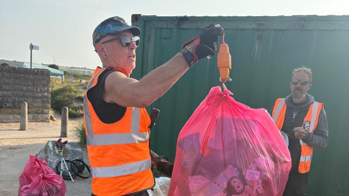 A man wearing a black t shirt and cap, sunglasses and an orange high viz vest. He is holding up a full red rubbish bag which is attached to a weighing scale. In the background are more rubbish bags and another man in orange high viz.