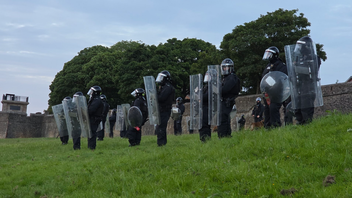 Shows riot police dressed in black with helmets and riot shields standing on a grass verge with Derry walls and trees in the background.
