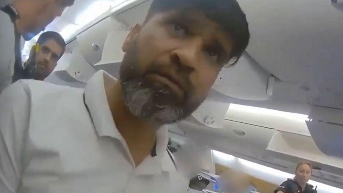 A man with dark hair and a dark and grey beard looking just above the camera. He is wearing a white polo shirt and is on an aeroplane