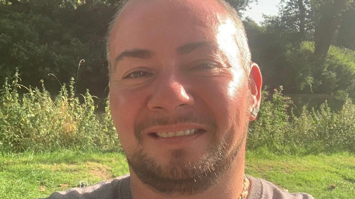 A closeup photo of a man with very short brown hair and a goatee beard smiling at the camera in a field. 