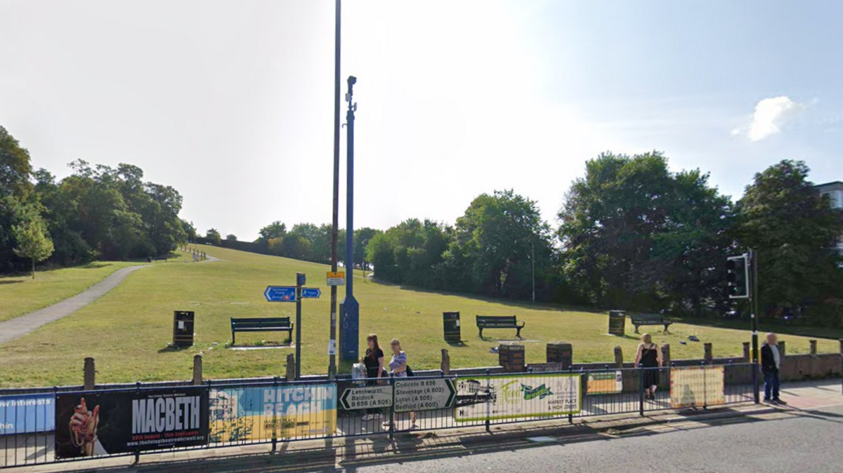 A view of a steep hill taken from a road. There are road signs on railings at the bottom of the hill. There is a CCTV camera at the bottom of the hill as well as benches and bins and people walking on the pavement.