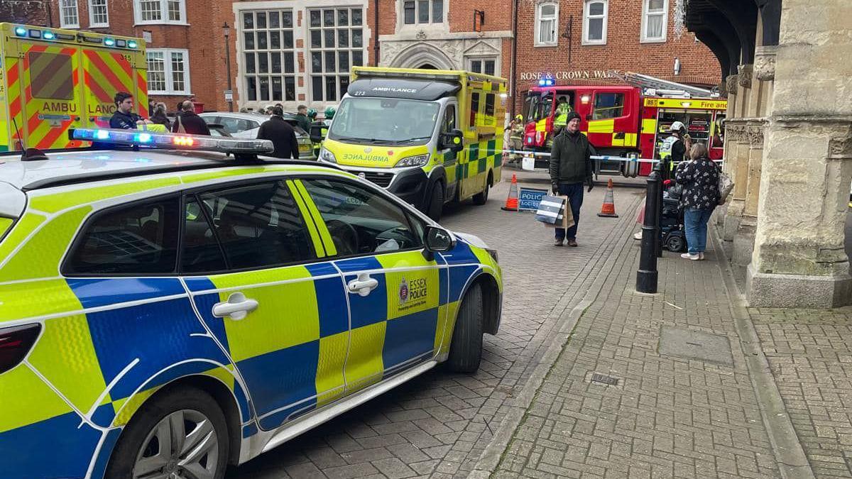 An Essex police car, two ambulances, fire engines in Market Place, Saffron Walden. The area has been cordoned off and a number of people are standing by it. You can see historic brick buildings surrounding the area.