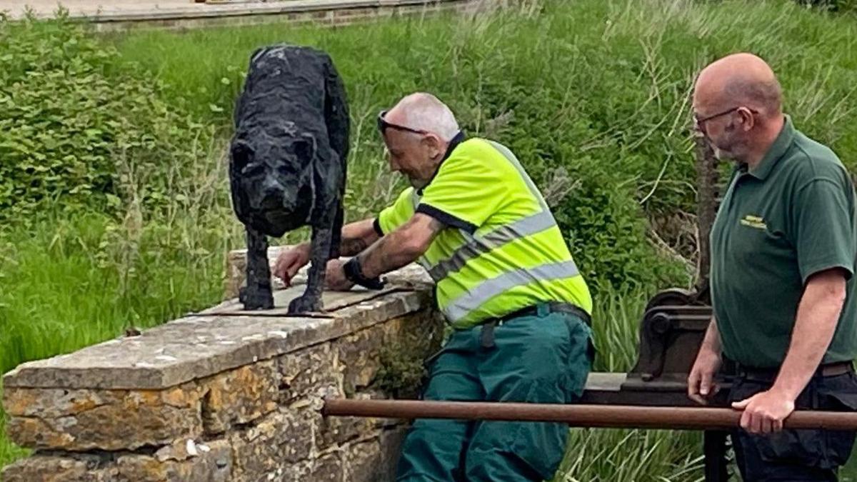 Two men removing the dog sculpture from the top of the wall next to the weir. Both are wearing green and one is wearing a hi-vis vest.