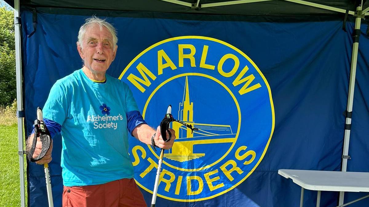 Mr Standring wears a bright blue Alzheimer's Society top, and smiles at the camera holding two walking poles, He is in front of a branded backdrop with a logo that says 'Marlow Striders'