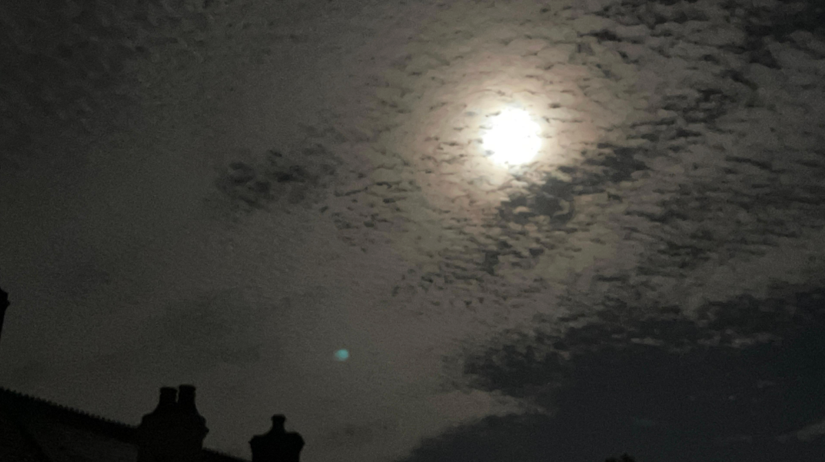 The moon is breaking through the clouds lighting up the night sky over some houses. It's bright and is lighting up the roof tops of houses.
