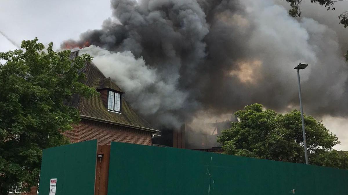 Smoke billowing from Henderson Hall in Newcastle. Flames can be seen just above the roof of the building on its far side. The building is made of brown brick and has a dormer window in its roof. It is located behind green solid wooden panels.