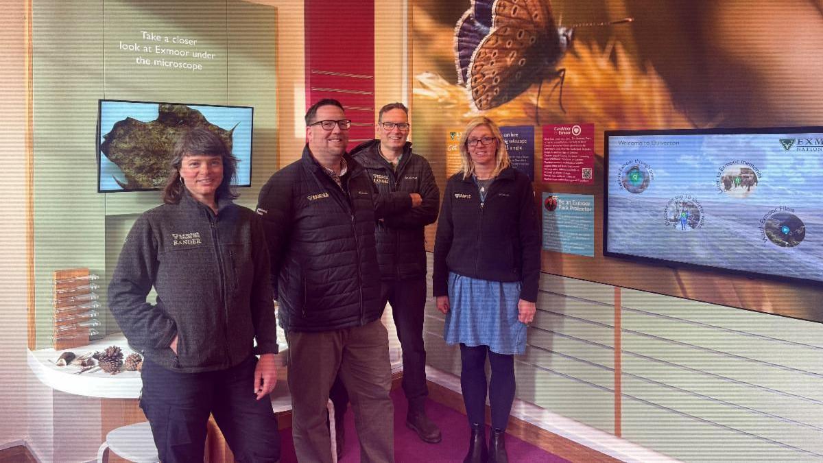 Four national park rangers with black zip-up jackets on. They are standing in the new headquarters in front of a large display wall which shows a butterfly on a flower and a TV screen. Behind them is a table full of natural materials collected from the national park. 