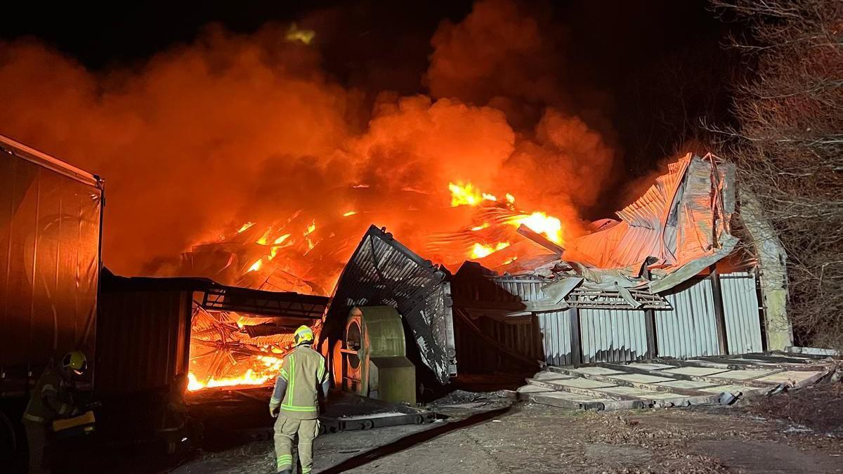 A firefighter stood watching a large blaze which is lighting up the night sky in red and orange. There are huge plumes of smoke billowing from it. The former hangar is falling apart due to the blaze